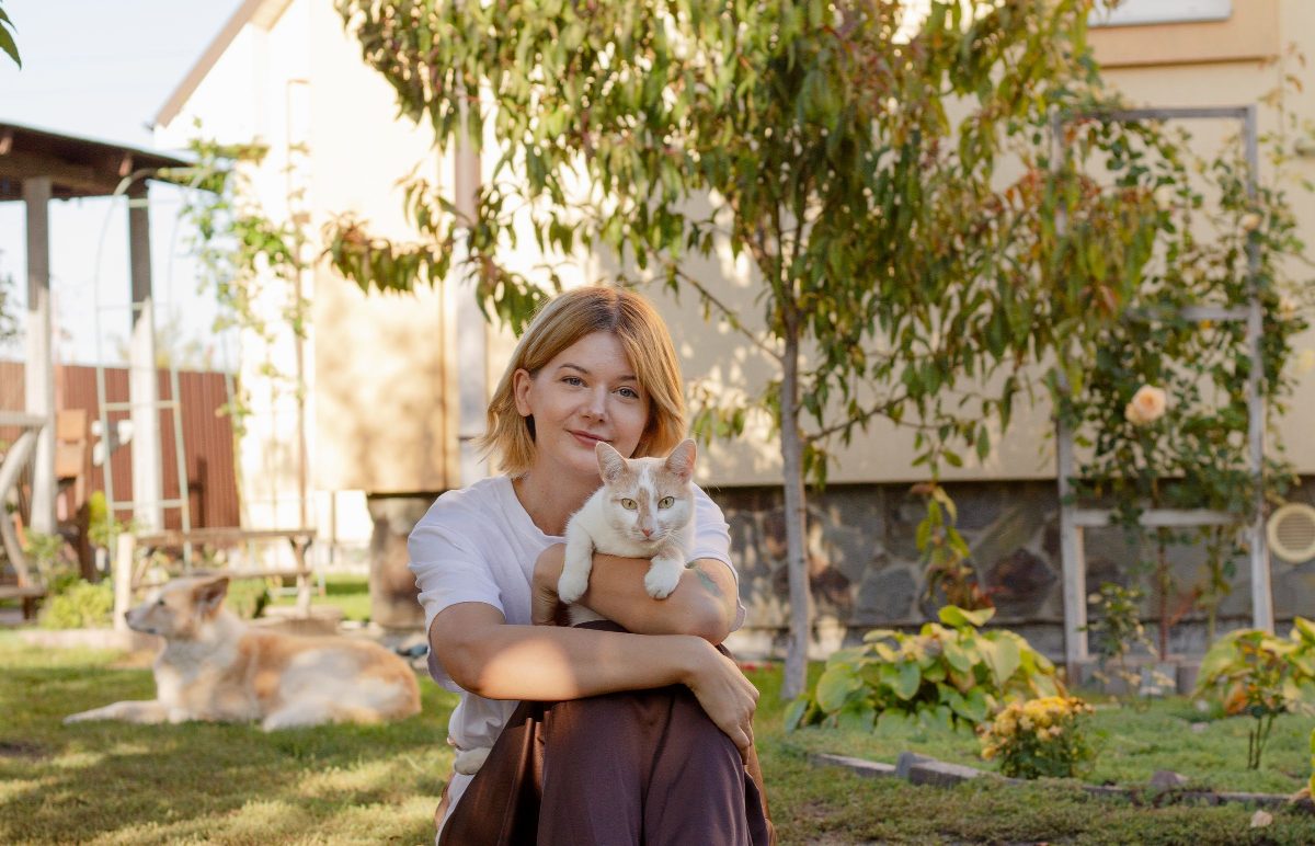 A pet sitter in a garden holding a cat, another cat resting on the grass